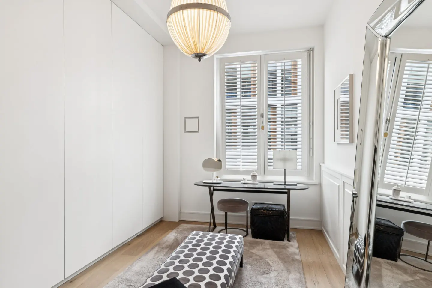 Bright, white room with a desk by a window, a polka-dot bench, and a large mirror. A globe-shaped light fixture hangs from the ceiling.