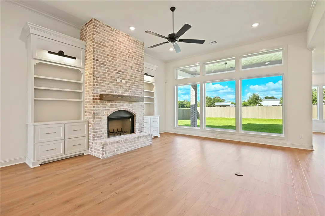 Living room with light wood floors, a brick fireplace, built-in white shelving, and large windows overlooking a backyard.