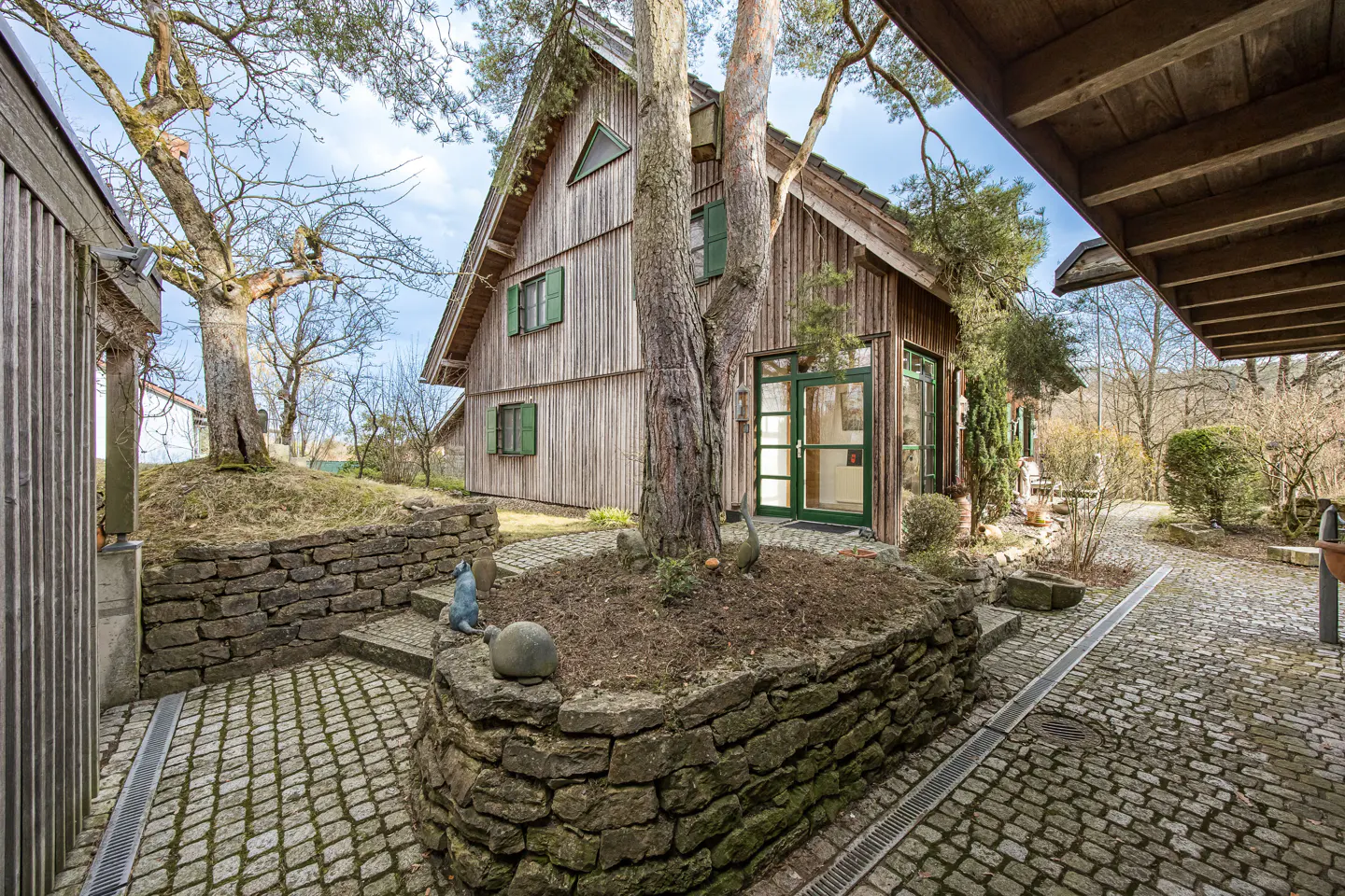 Exterior view of a wood-sided house with green trim, a stone patio, and a tree in the front yard.
