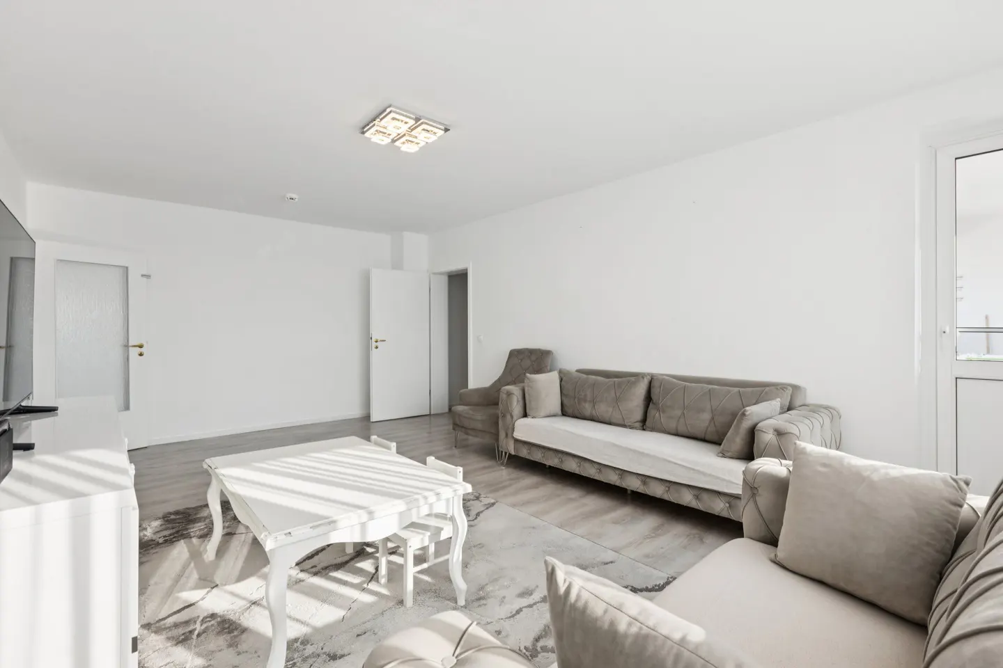 Bright living room with white walls, light wood floors, and neutral-toned sofas. A white table and chairs sit on a patterned rug.