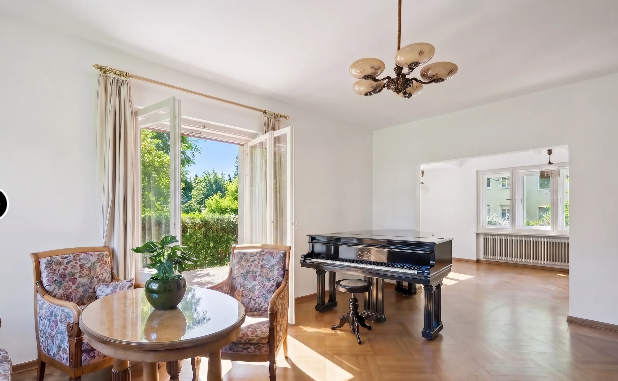 Bright living room with a black grand piano, a round table with two floral chairs, and open doors to a sunny garden. [AI-generated text]