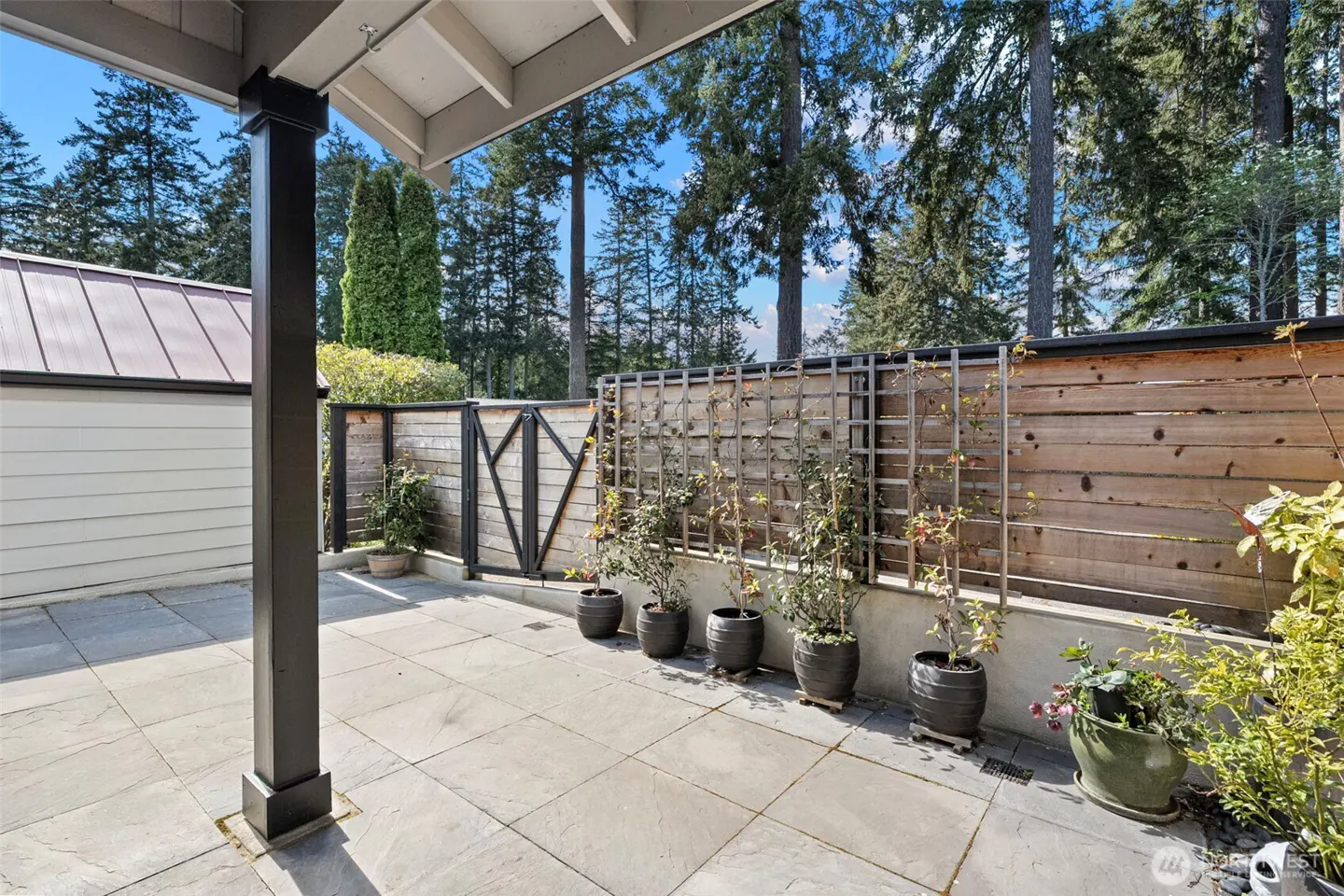 Outdoor patio with stone pavers, a dark pillar, and a wooden fence with potted plants. Tall green trees are visible in the background.