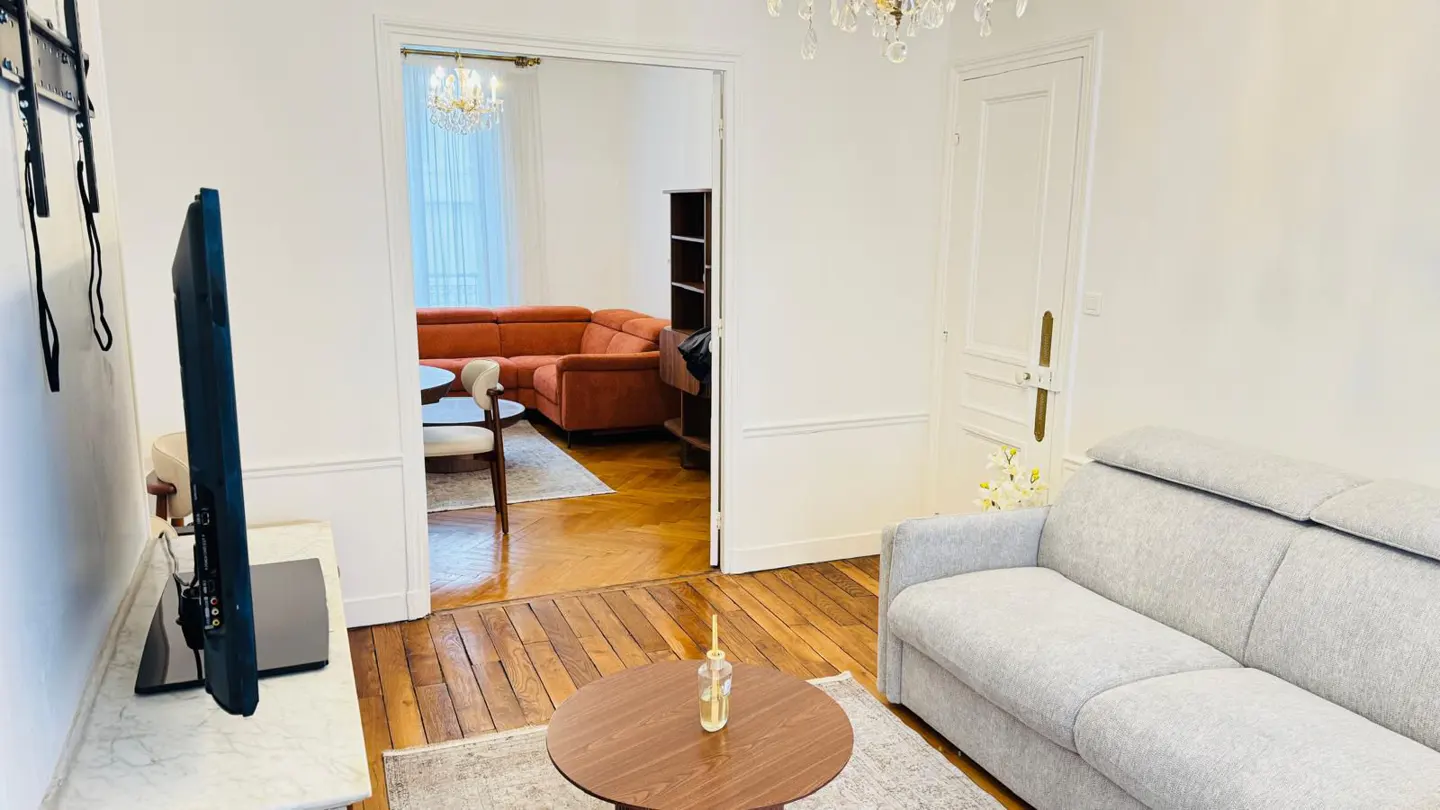 Living room with a gray sofa, a round wooden table, and a TV. An orange sofa is visible through a doorway.