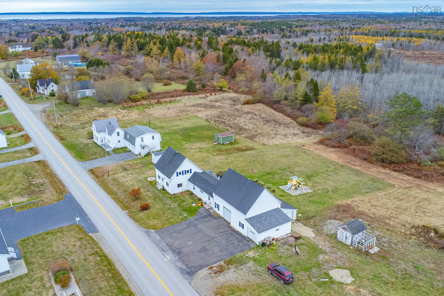 Aerial view of a white house with a gray roof, a playground, and a red car in a rural area with trees and a road.