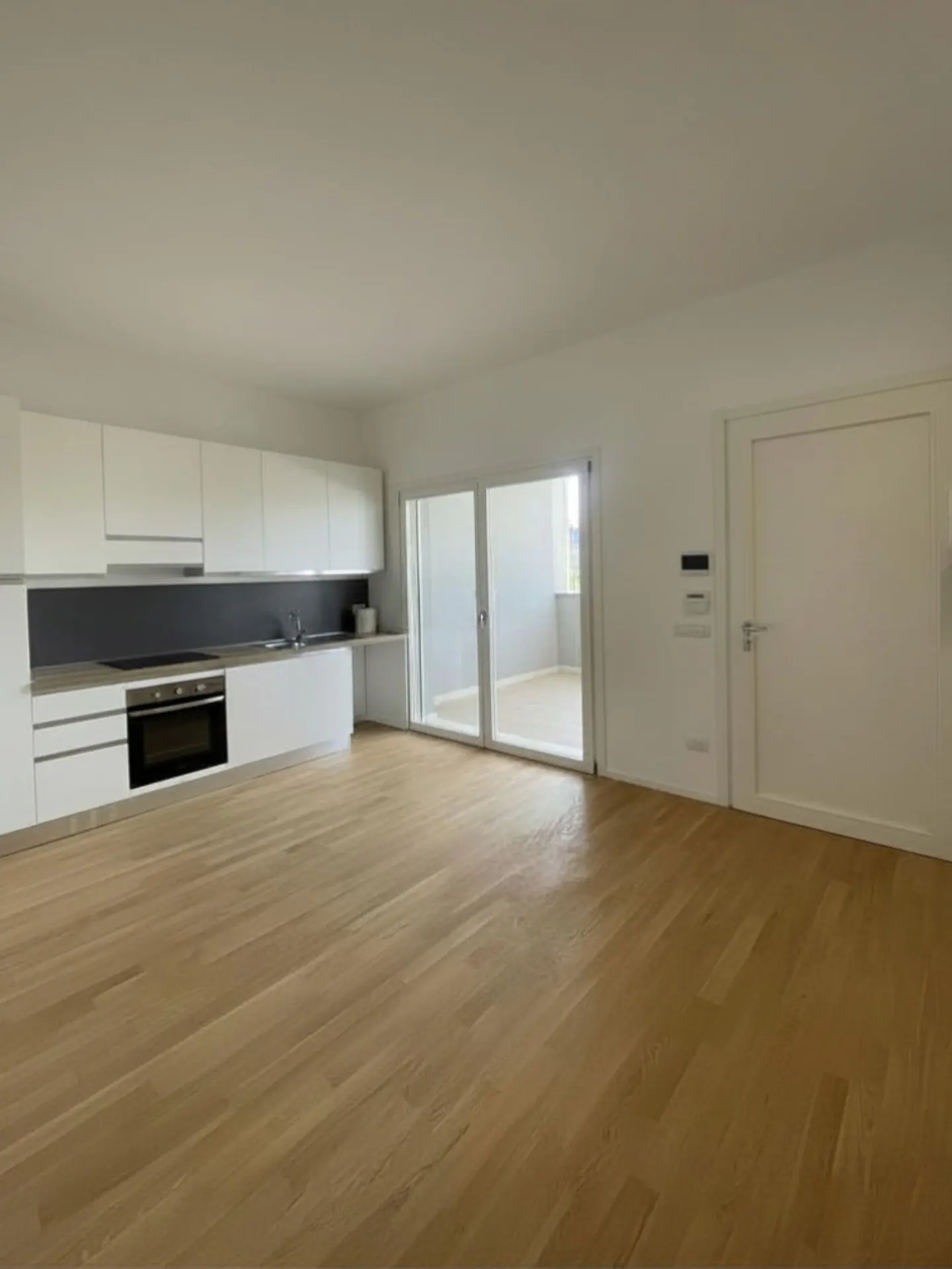 Bright, empty apartment with light wood floors, white walls, and a modern white kitchen. Sliding glass doors lead to a balcony.