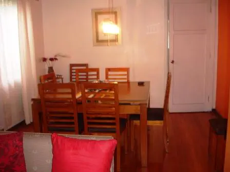 A dining room with a wooden table and chairs, a white door, and a red pillow on a couch.