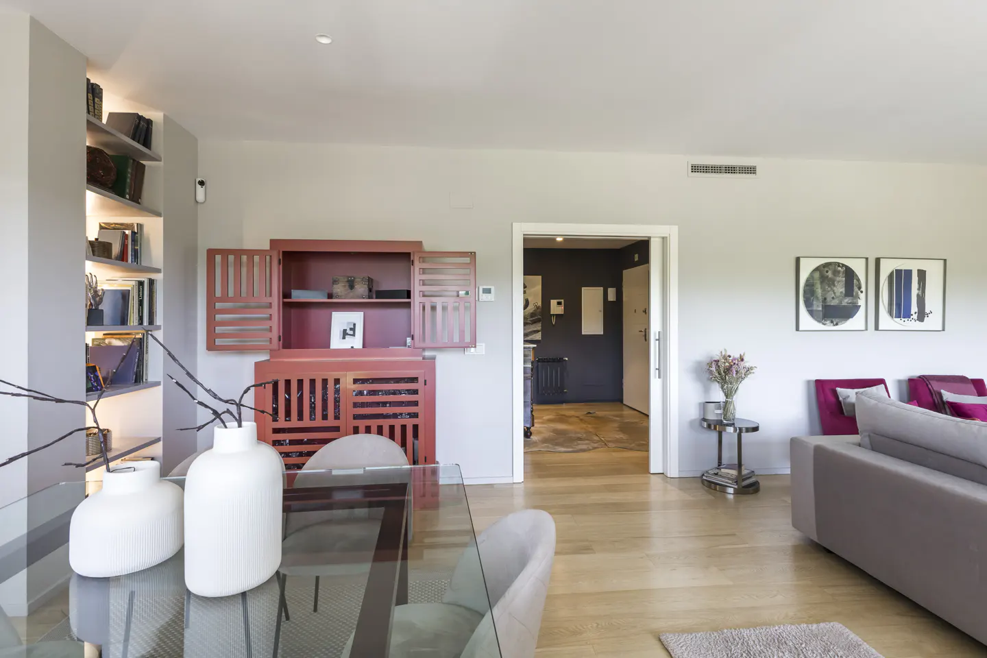 Modern living room with a glass dining table, red cabinet, built-in shelves, and a grey sofa on light wood flooring.