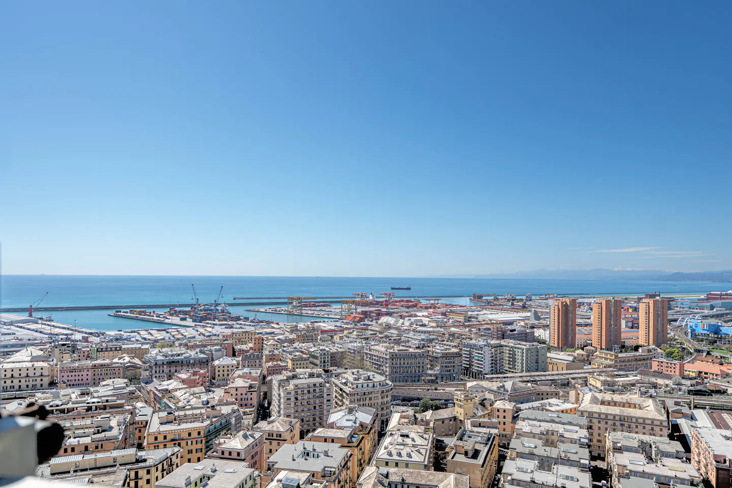 Cityscape view of Genoa, Italy, with the port and sea in the background under a clear blue sky. Buildings are mostly light-colored.
