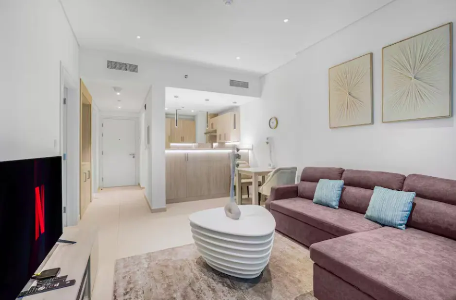 A modern apartment living room with a purple sofa, white coffee table, and open kitchen. Netflix is on the TV.