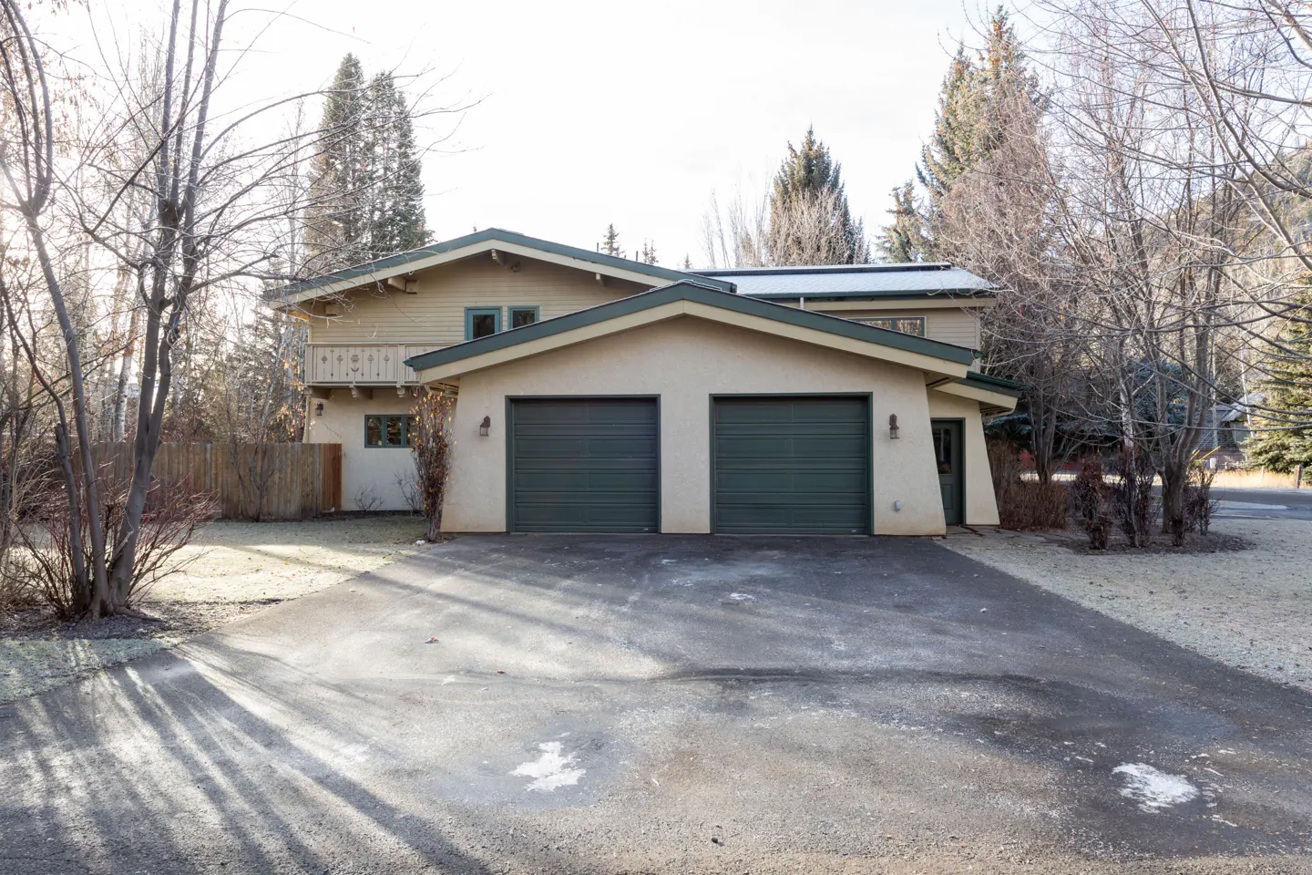Two-story house with a two-car garage and a paved driveway. The house is beige with green trim and garage doors. Trees surround the house.