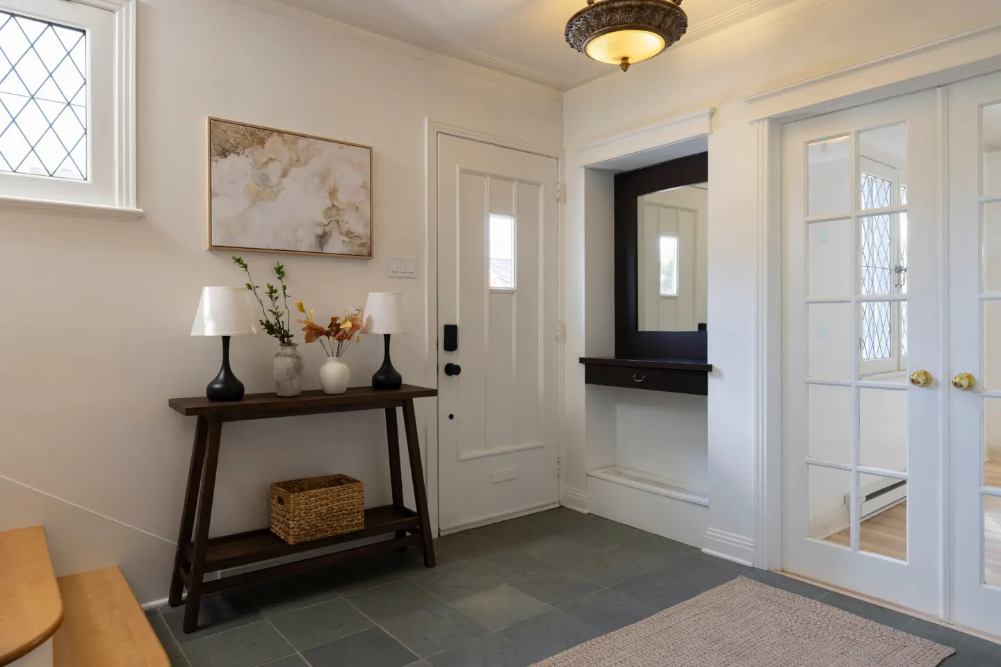 Entryway with white walls, gray tile floor, and a dark wood console table with lamps and flowers. A white door and glass paneled doors are visible.