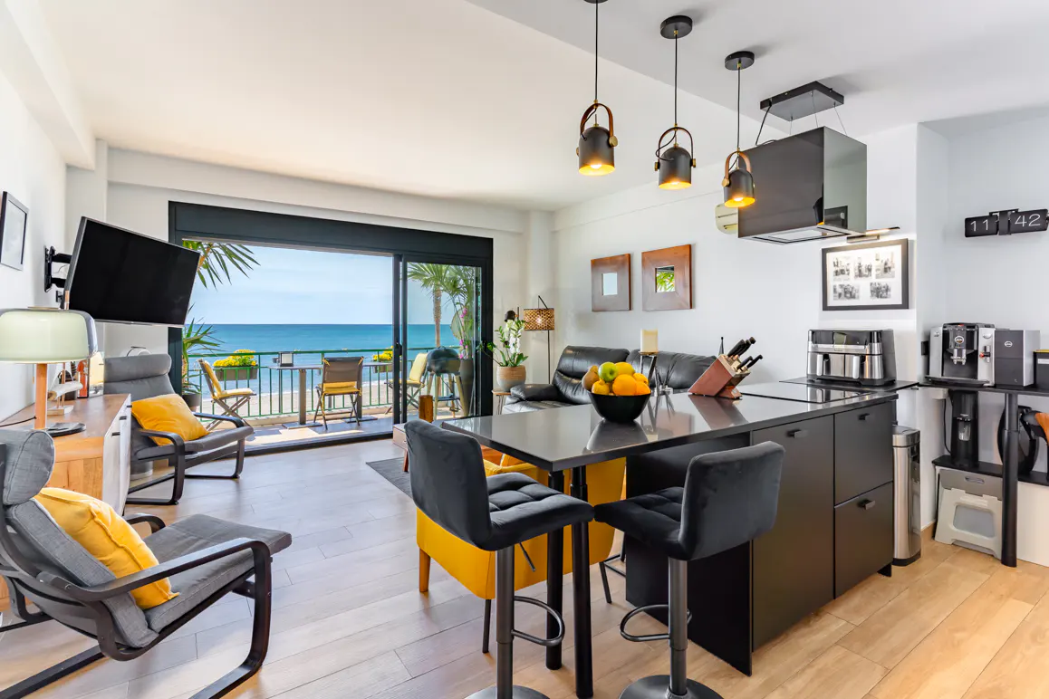 Modern open-plan apartment with a kitchen island, black bar stools, living area, and a balcony overlooking the ocean.