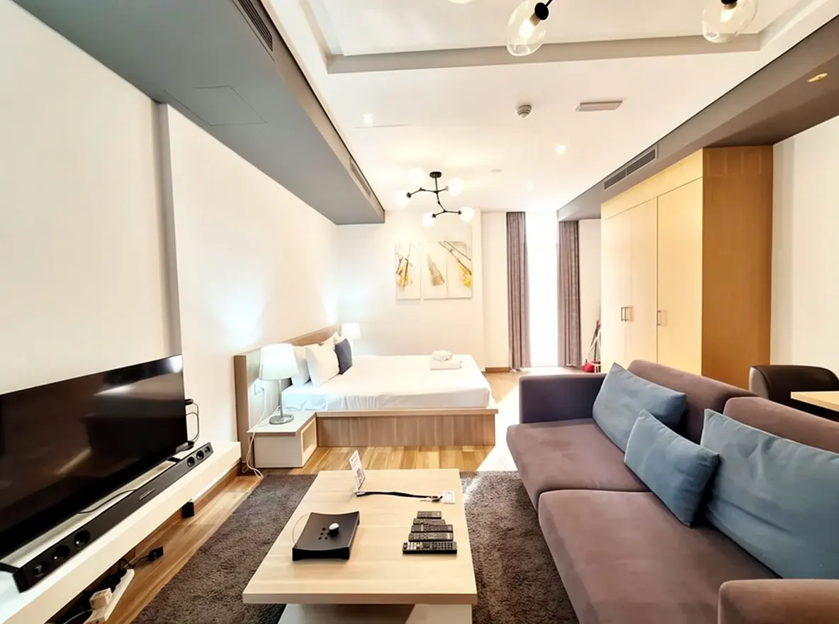 A bright studio apartment with a bed, sofa, TV, and coffee table. The walls are white, and the floor is wood.