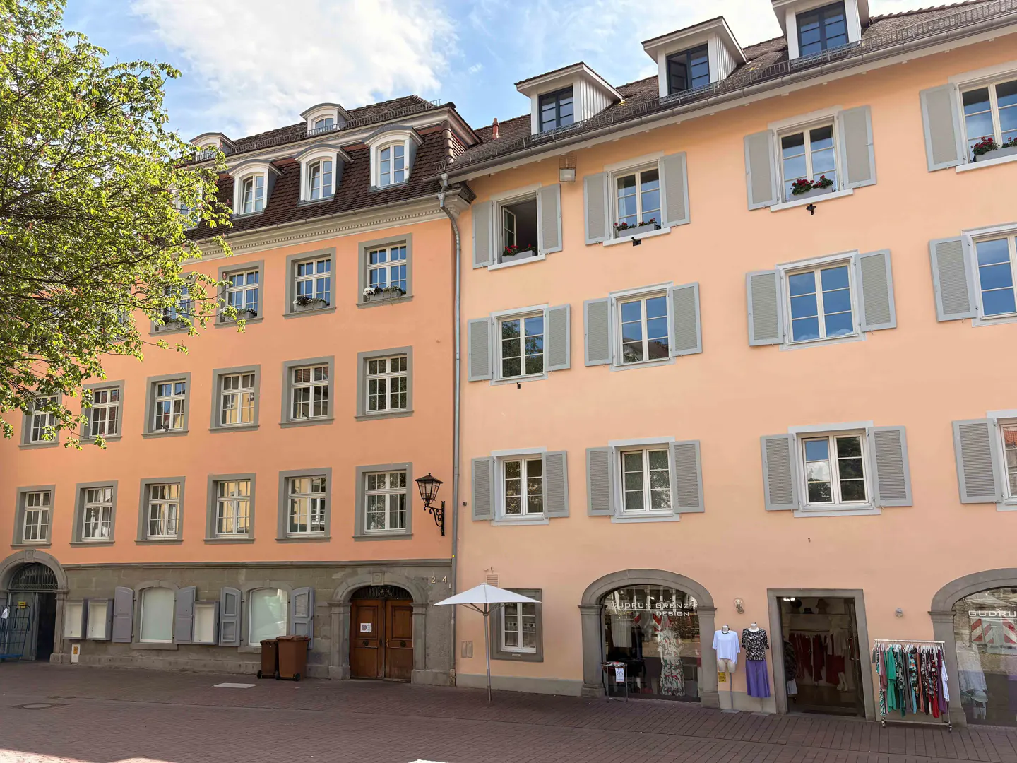 Peach-colored historic buildings with grey shutters and arched ground-floor storefronts on a sunny town square.