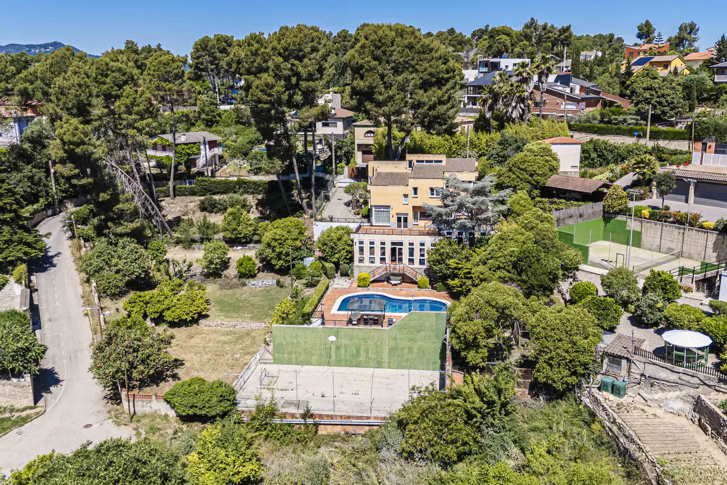 Aerial view of a yellow house with a pool, surrounded by trees and other houses on a sunny day.