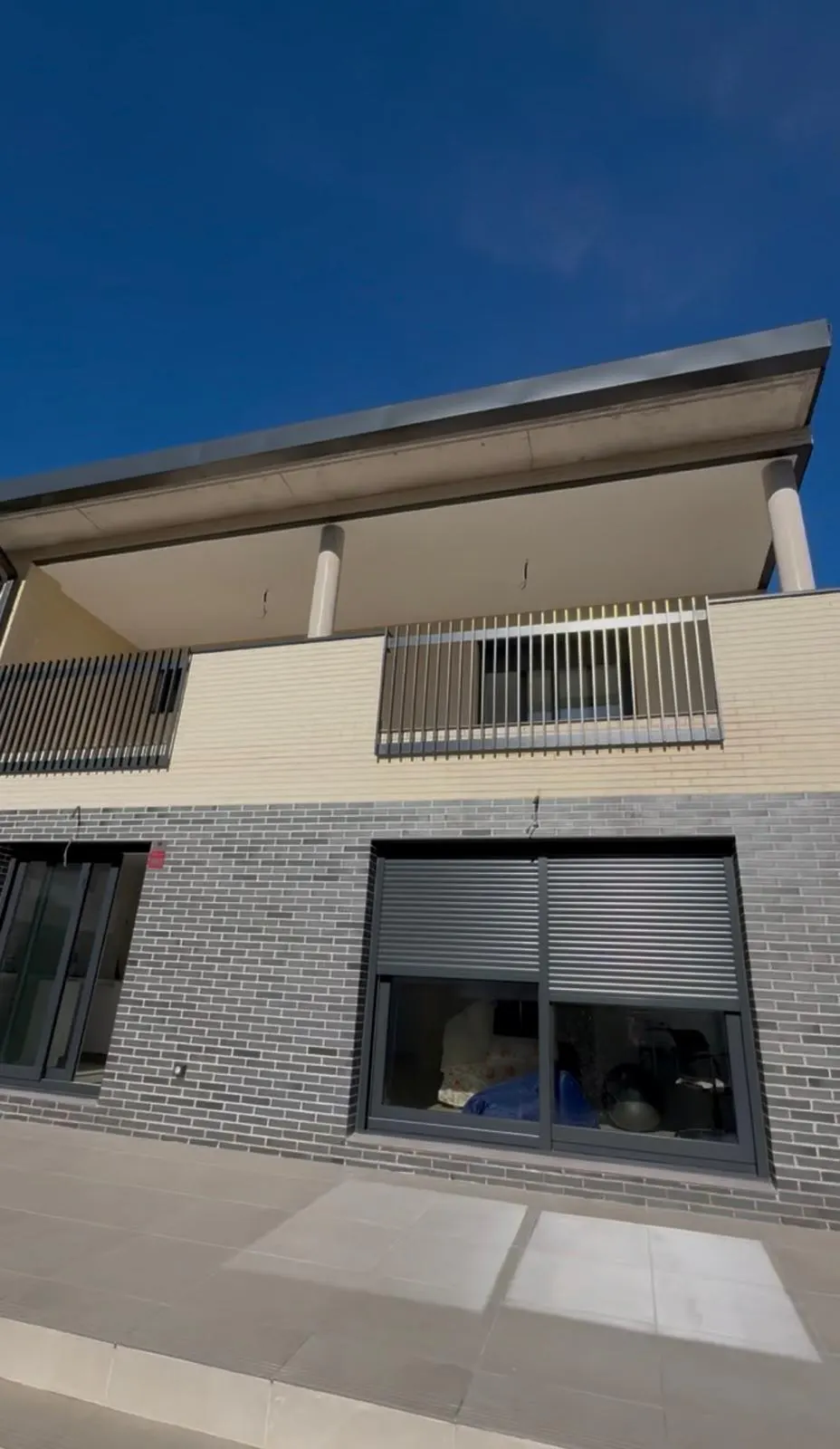 Exterior shot of a two-story building with gray brick, a balcony with metal railings, and a blue sky.