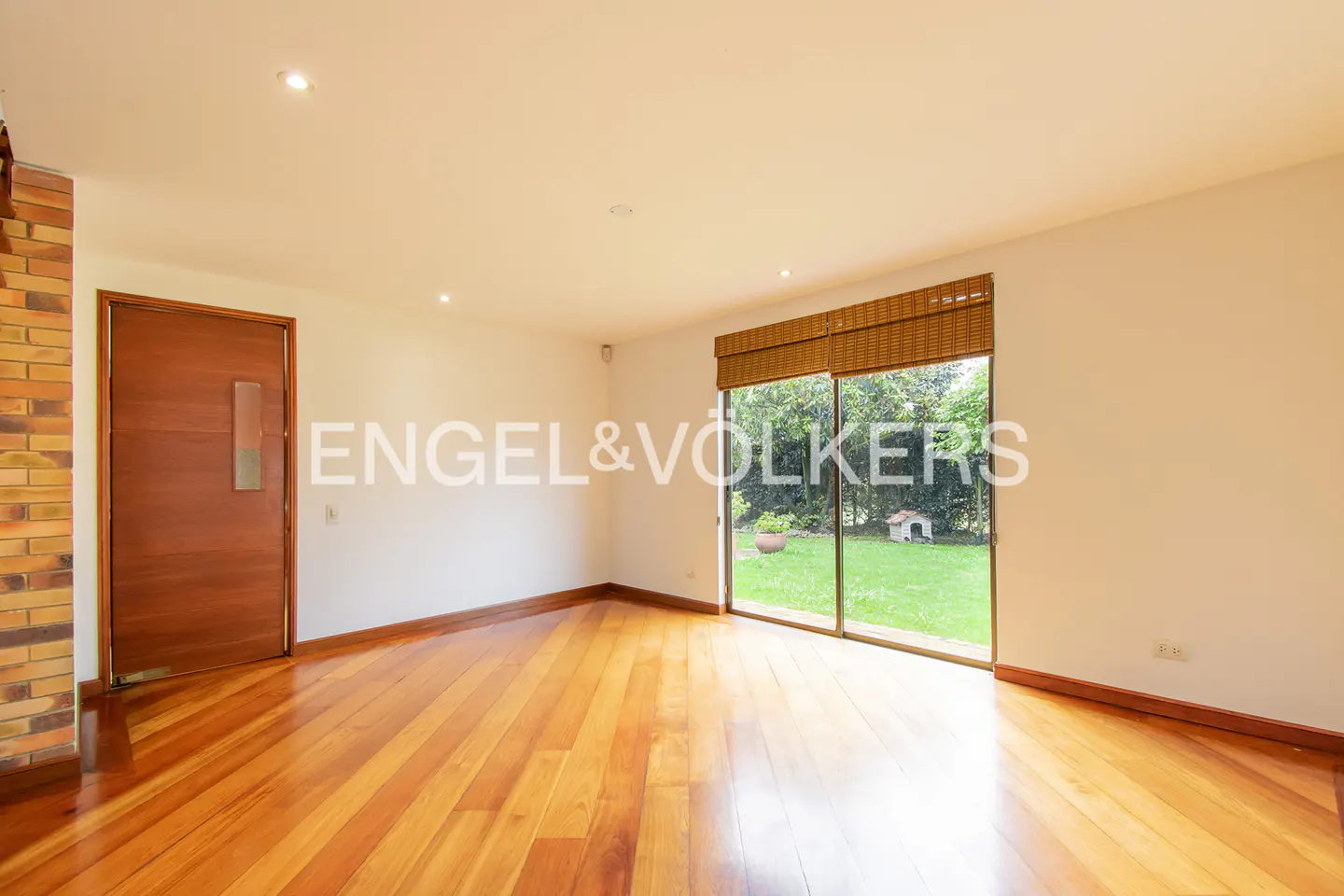 Empty room with hardwood floors, white walls, and a large window overlooking a green lawn. A wooden door is on the left.