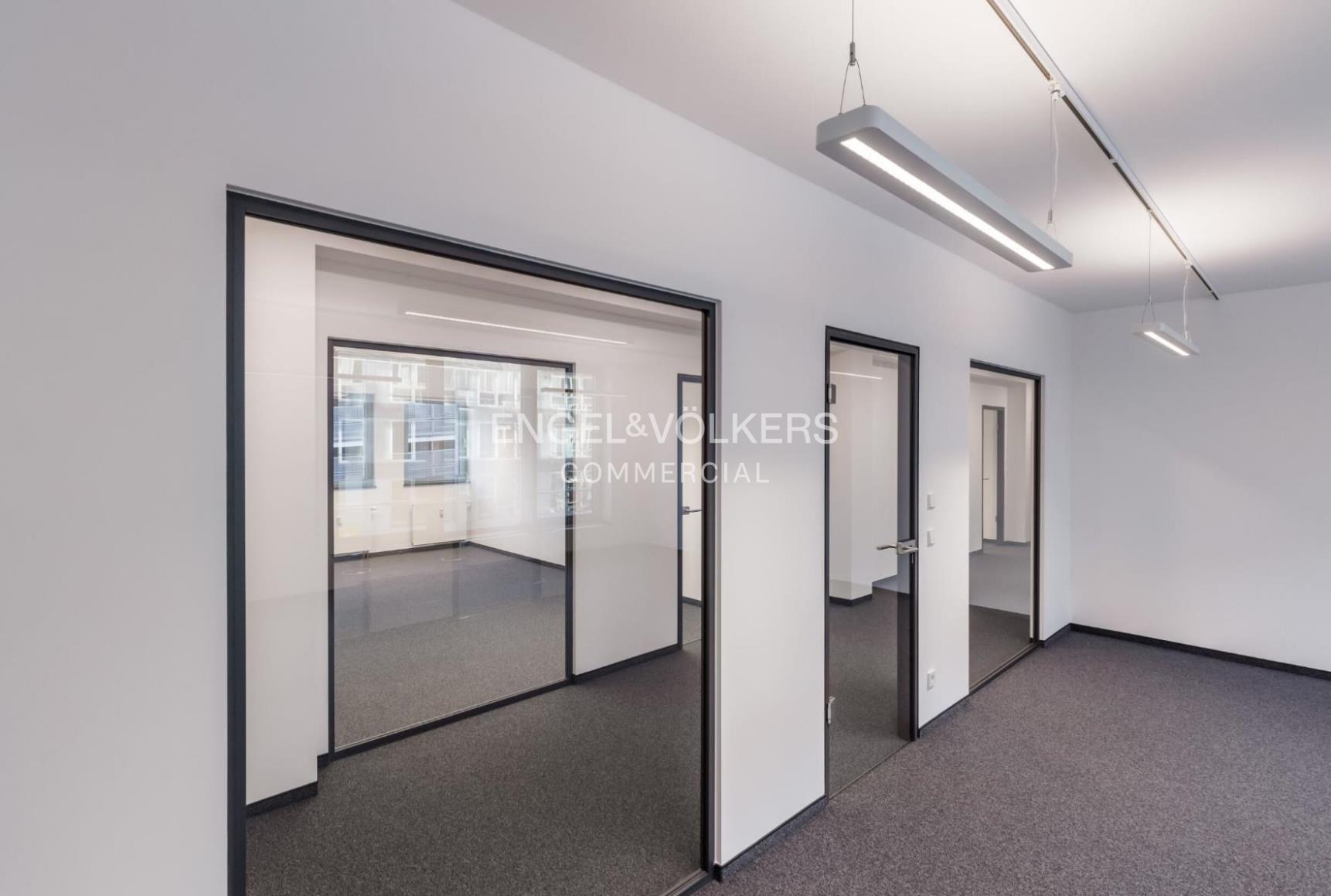 Modern office hallway with white walls, grey carpet, and glass-paneled doors leading to empty, well-lit workspaces.