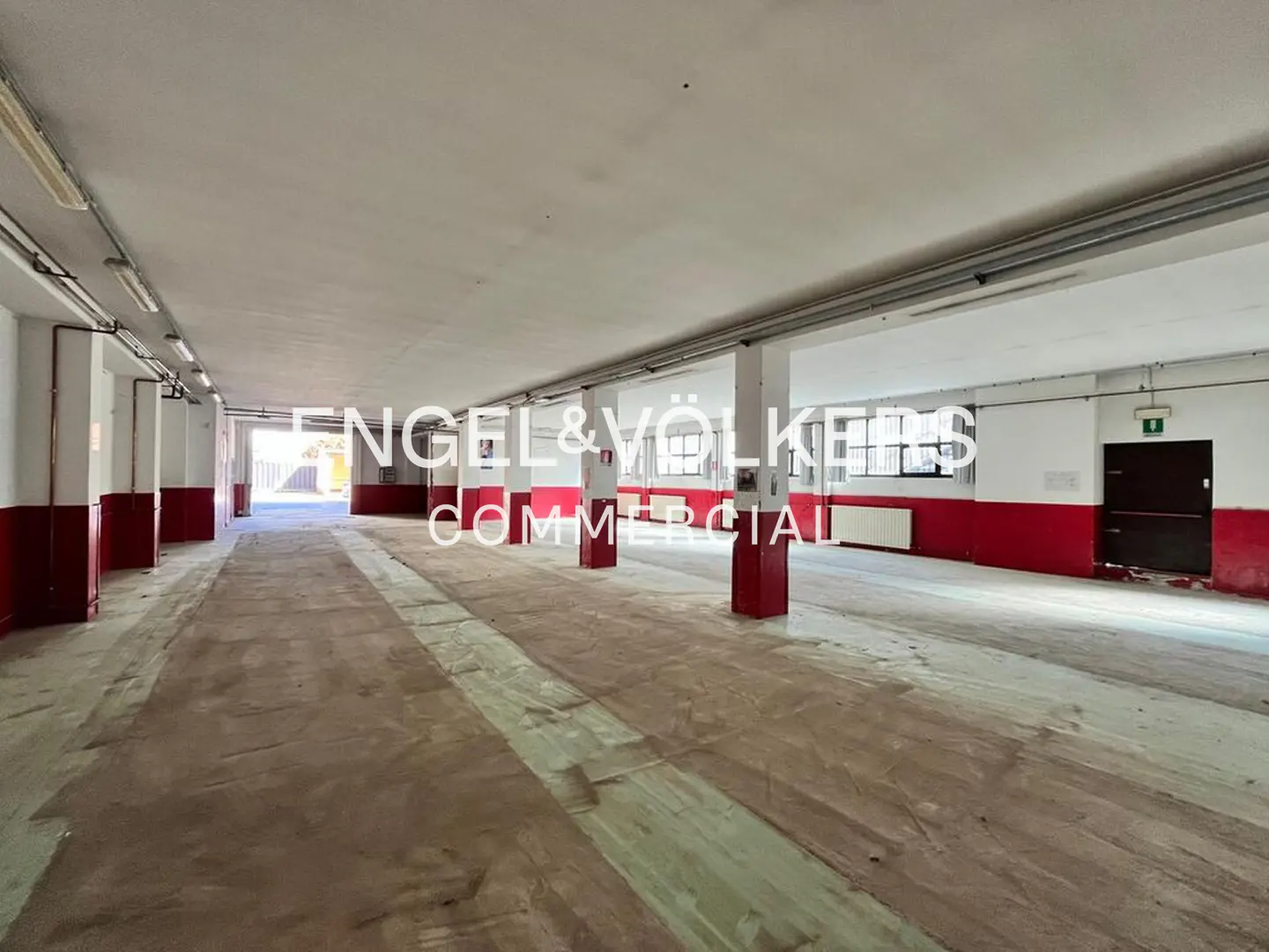Large, empty commercial space with white walls, red accents, and concrete floors. Columns support the ceiling. Engel & Völkers logo overlay.
