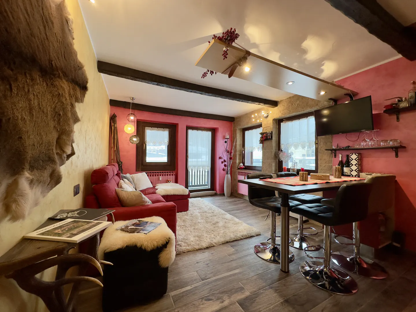 Cozy living room with red sofa, white rug, and dining table with black chairs. Walls are pink and yellow, with wooden beams on the ceiling.