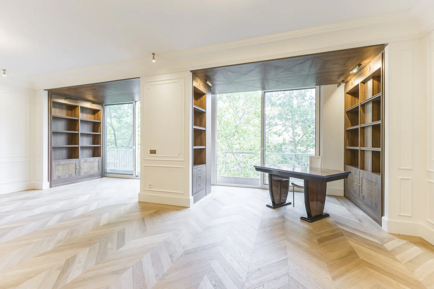 Bright room with herringbone wood floors, built-in bookshelves, and a dark wood desk by a large window.