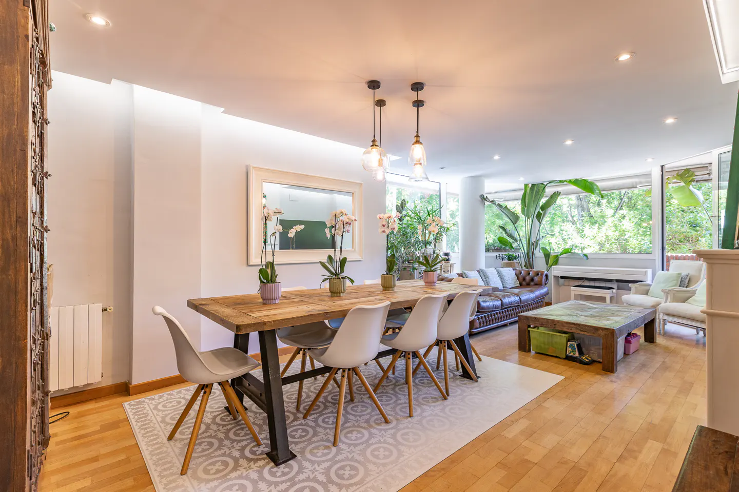 Bright dining area with a rustic wooden table, white chairs, orchid centerpieces, and an open-plan living room beyond.