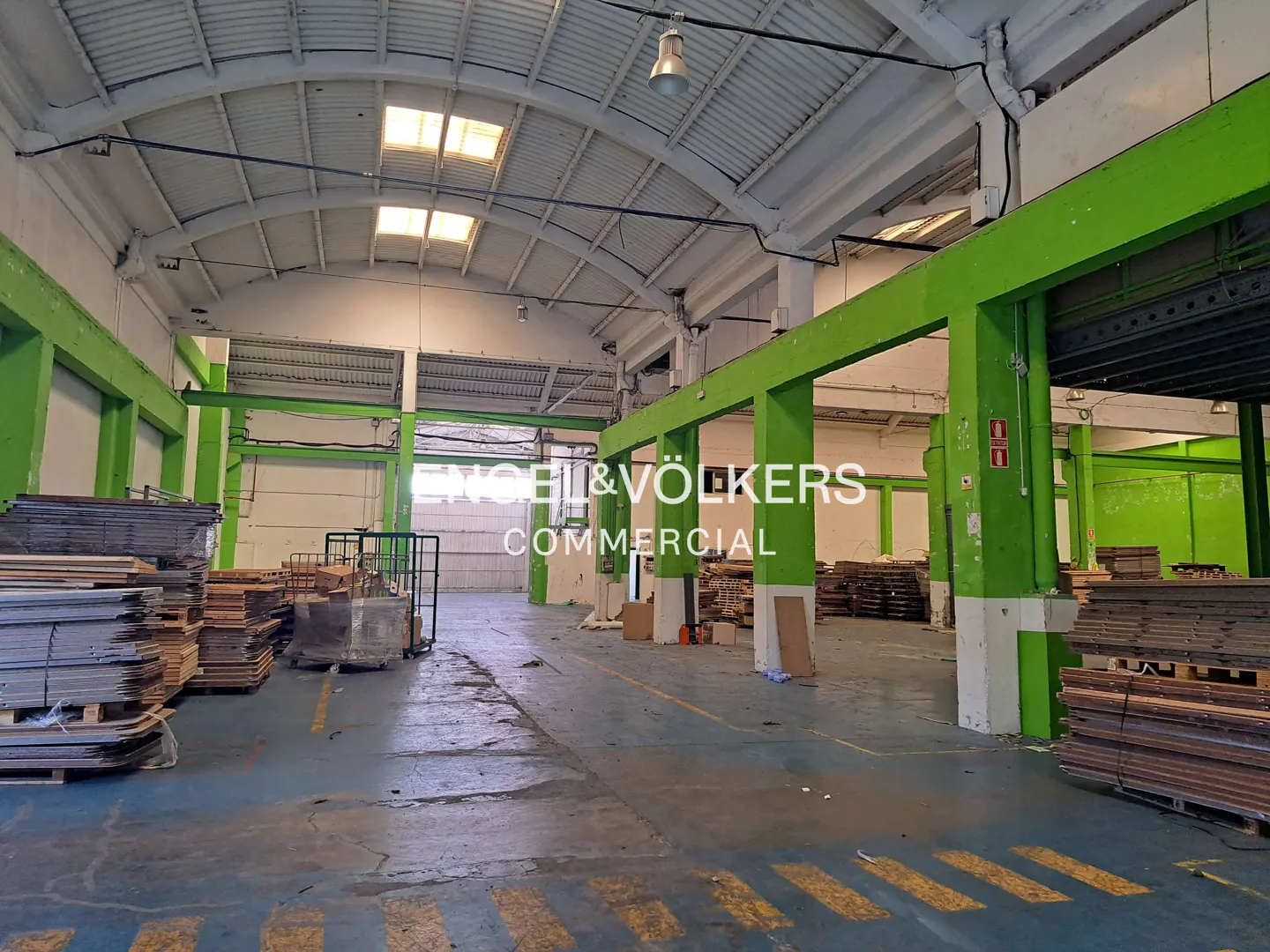 An interior shot of a large warehouse with green support beams and stacks of wooden pallets.
