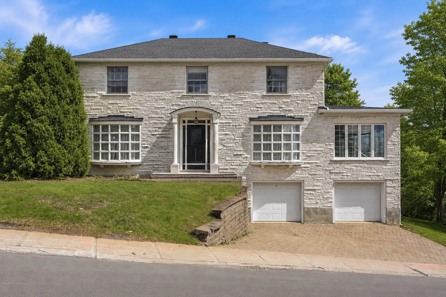 Two-story white stone house with a black roof, two garage doors, and a green lawn.