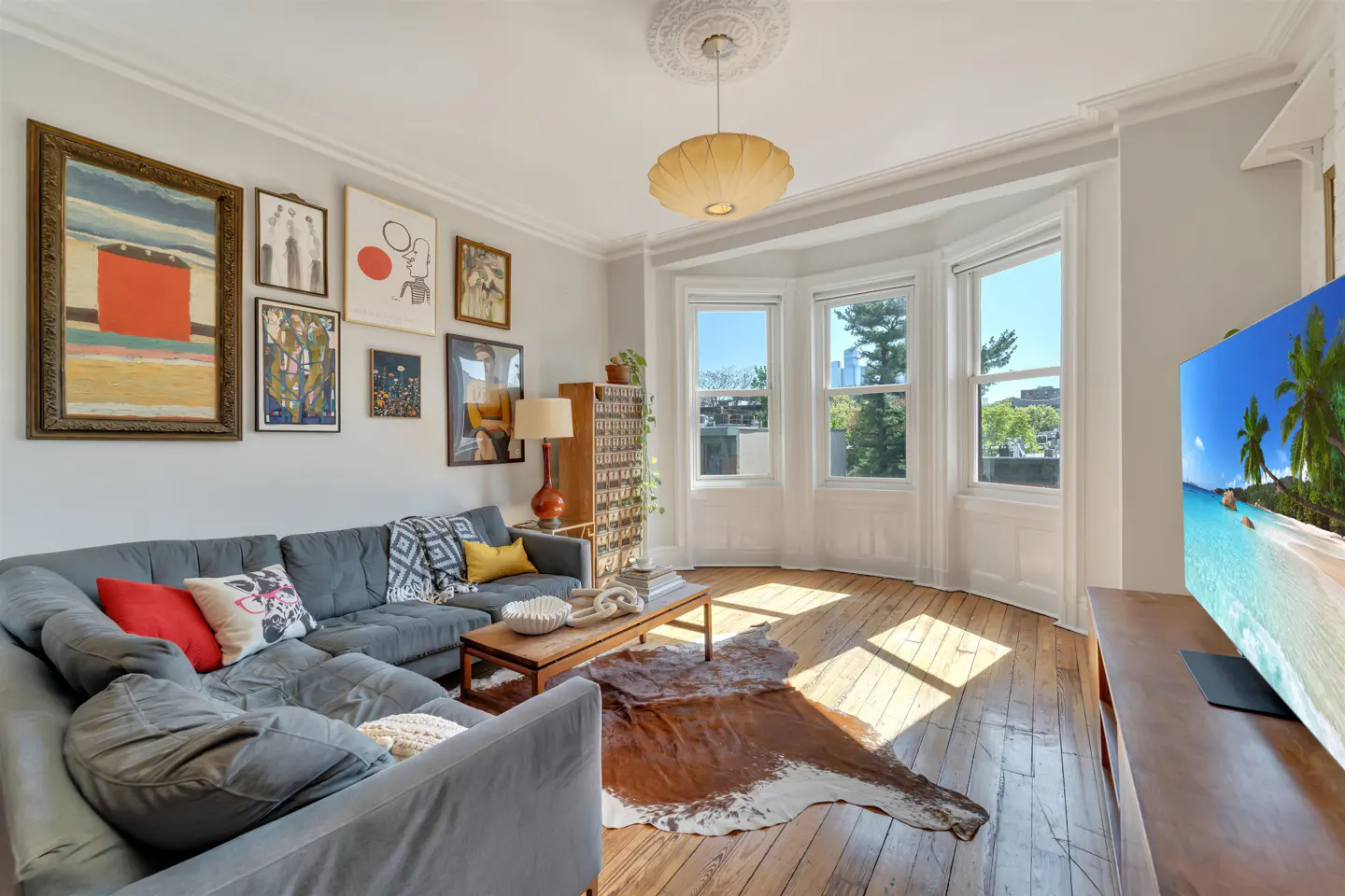 Living room with gray sectional sofa, wood floors, bay window, and gallery wall. A cowhide rug is on the floor.