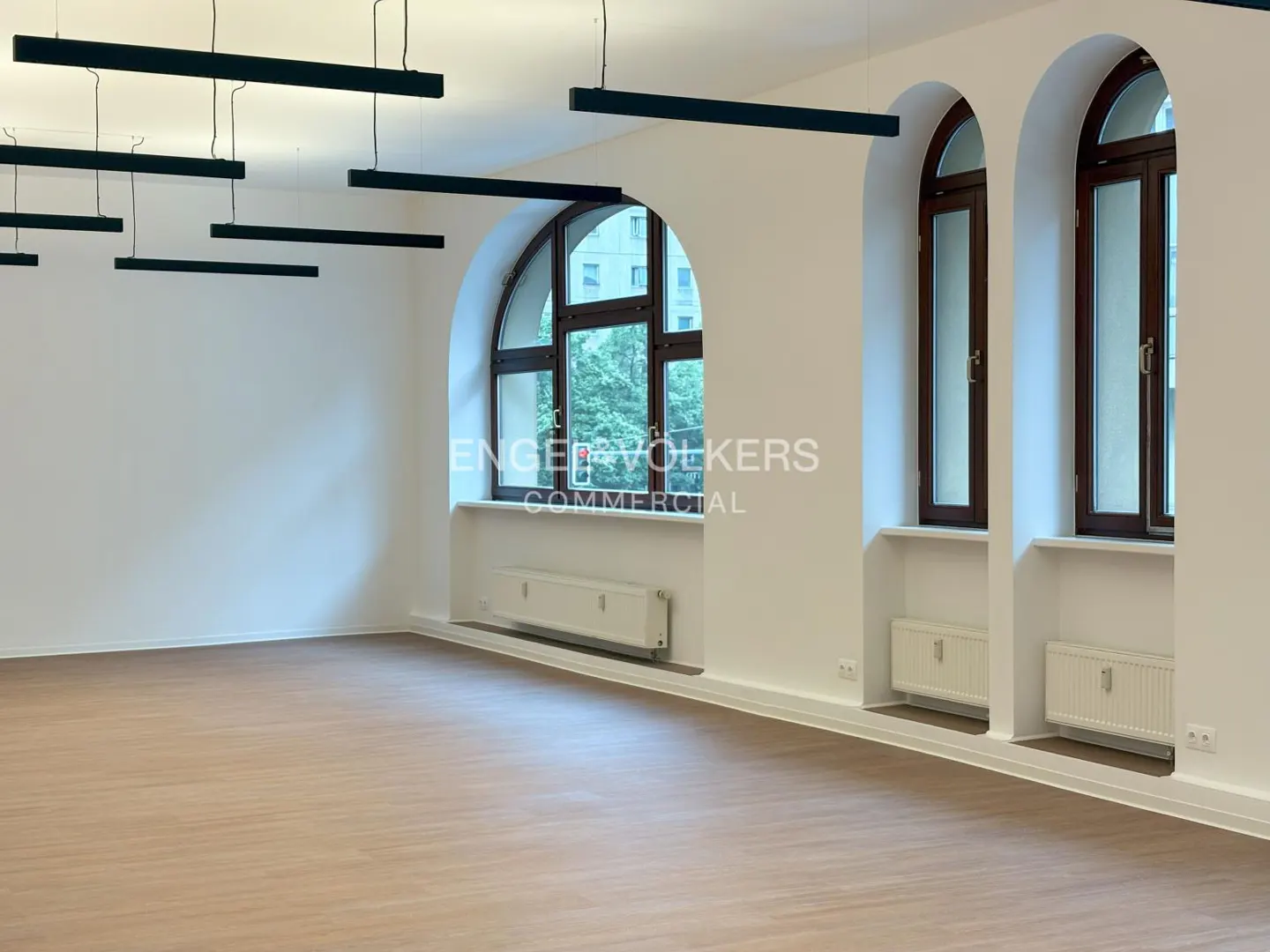 Bright, empty room with light wood floors, white walls, arched windows, and black linear pendant lights.