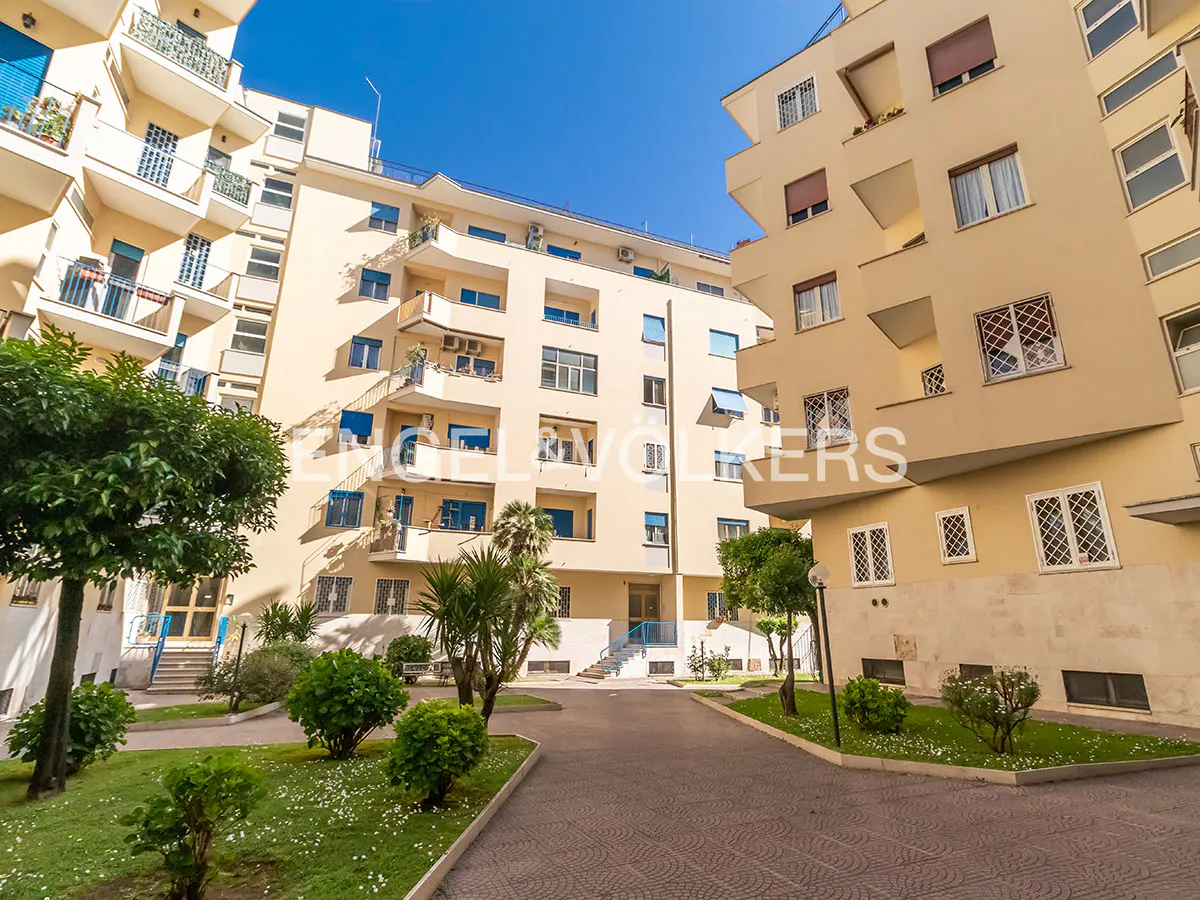 Exterior view of a multi-story apartment complex with balconies, green lawn, trees, and a paved walkway under a clear blue sky.