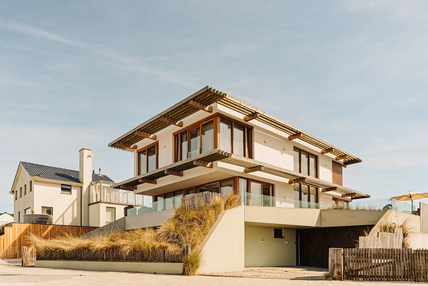 A modern, two-story white house with wood trim and large windows sits on a sandy lot with beach grass. A garage is built into the base of the house.