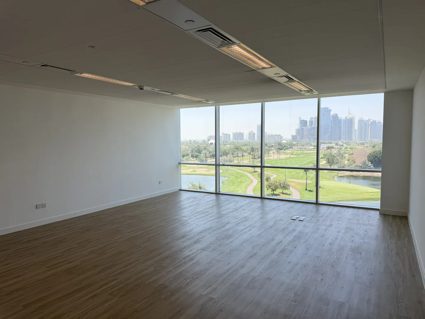 Empty office space with wood floors and white walls. Large windows overlook a golf course and city skyline.