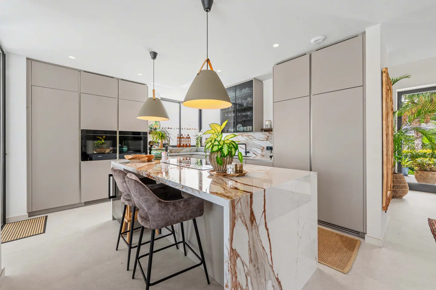 A modern kitchen with gray cabinets, a marble island with bar stools, and pendant lights. Plants add a touch of greenery.