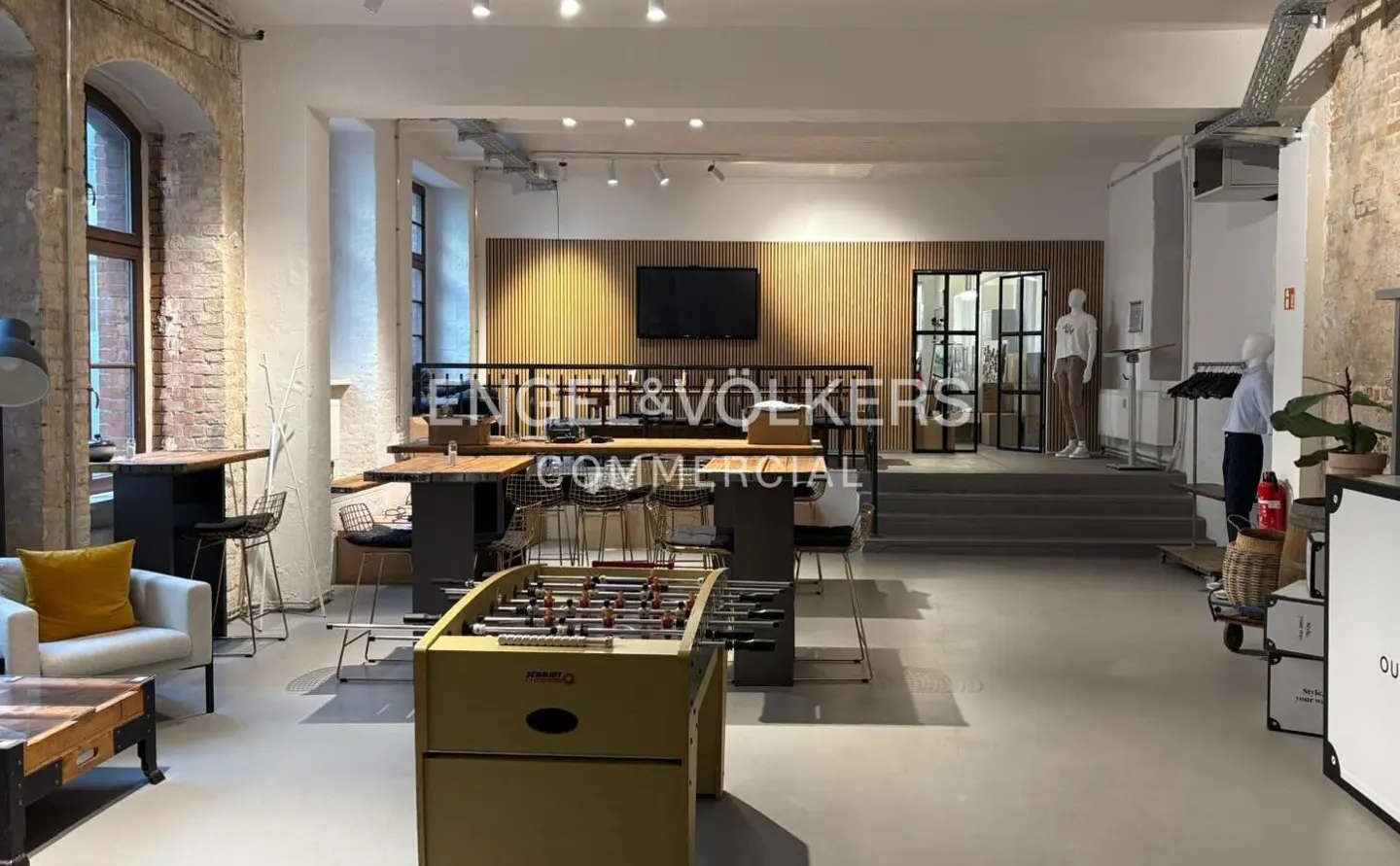 Bright, modern Engel & Völkers commercial space with a foosball table, lounge area, and high-top tables. Mannequins are visible in the background.