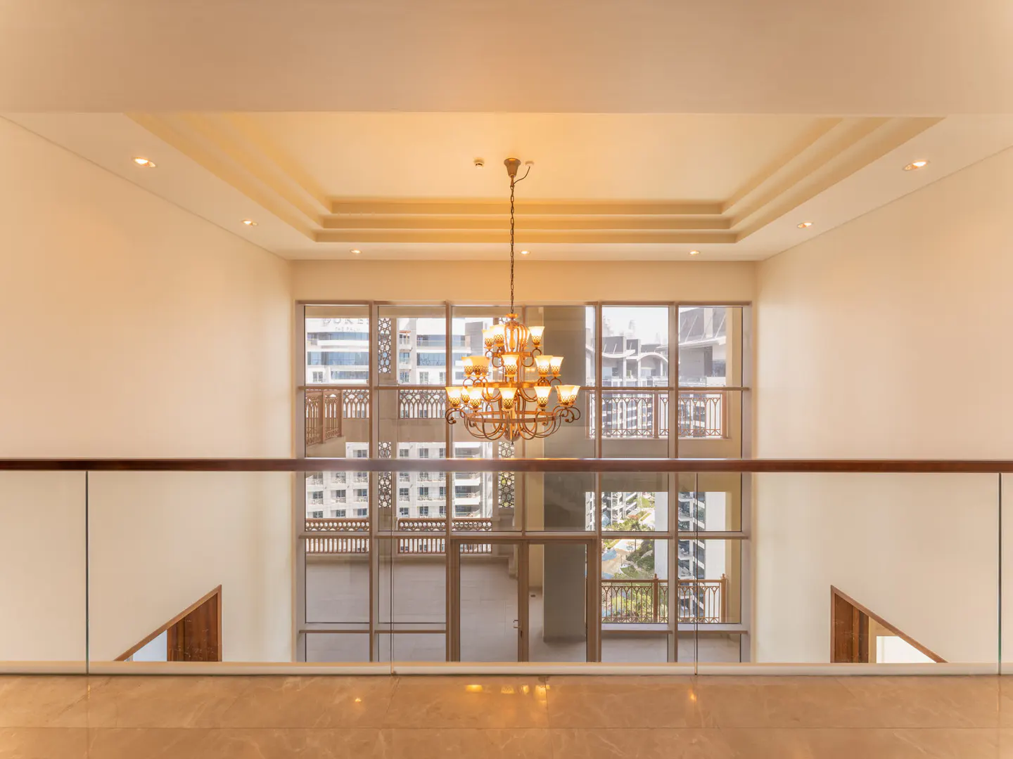 Interior view from a balcony with a chandelier, large windows, and a city view. Cream walls and marble floors.
