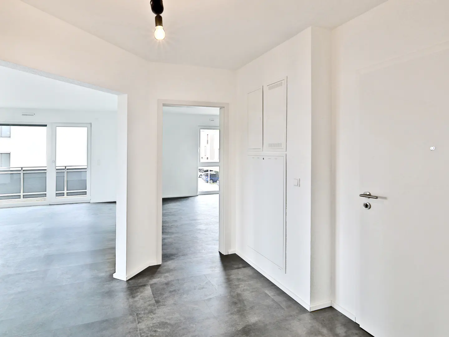 Bright, modern apartment interior with gray floors, white walls, and open doorways leading to other rooms.