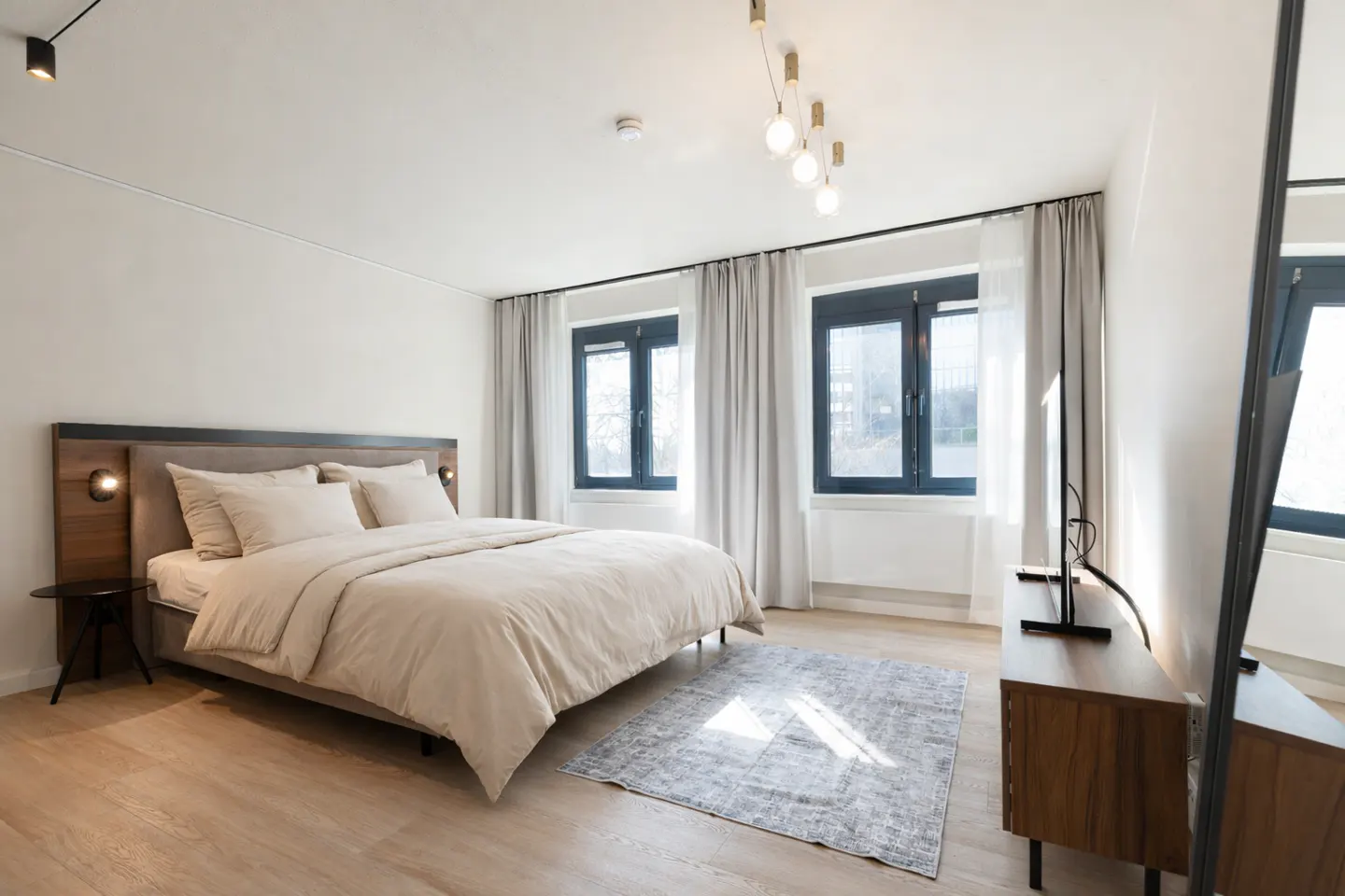 A bright bedroom with a queen bed, wood floors, and two windows with gray curtains. A gray rug is on the floor.