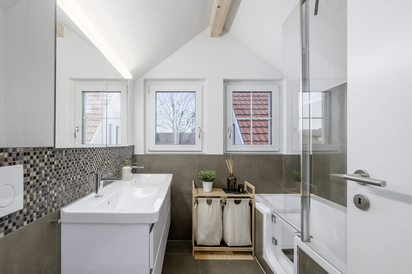 Bright bathroom with white walls, a sink, and a bathtub. Two windows offer a view of rooftops. A laundry basket stands near the tub.
