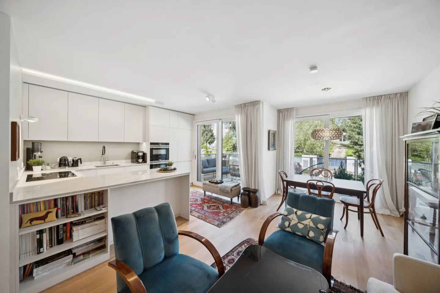 Bright, open-concept living space with white kitchen, dining table, and blue velvet armchairs. Sliding glass doors lead to a balcony.