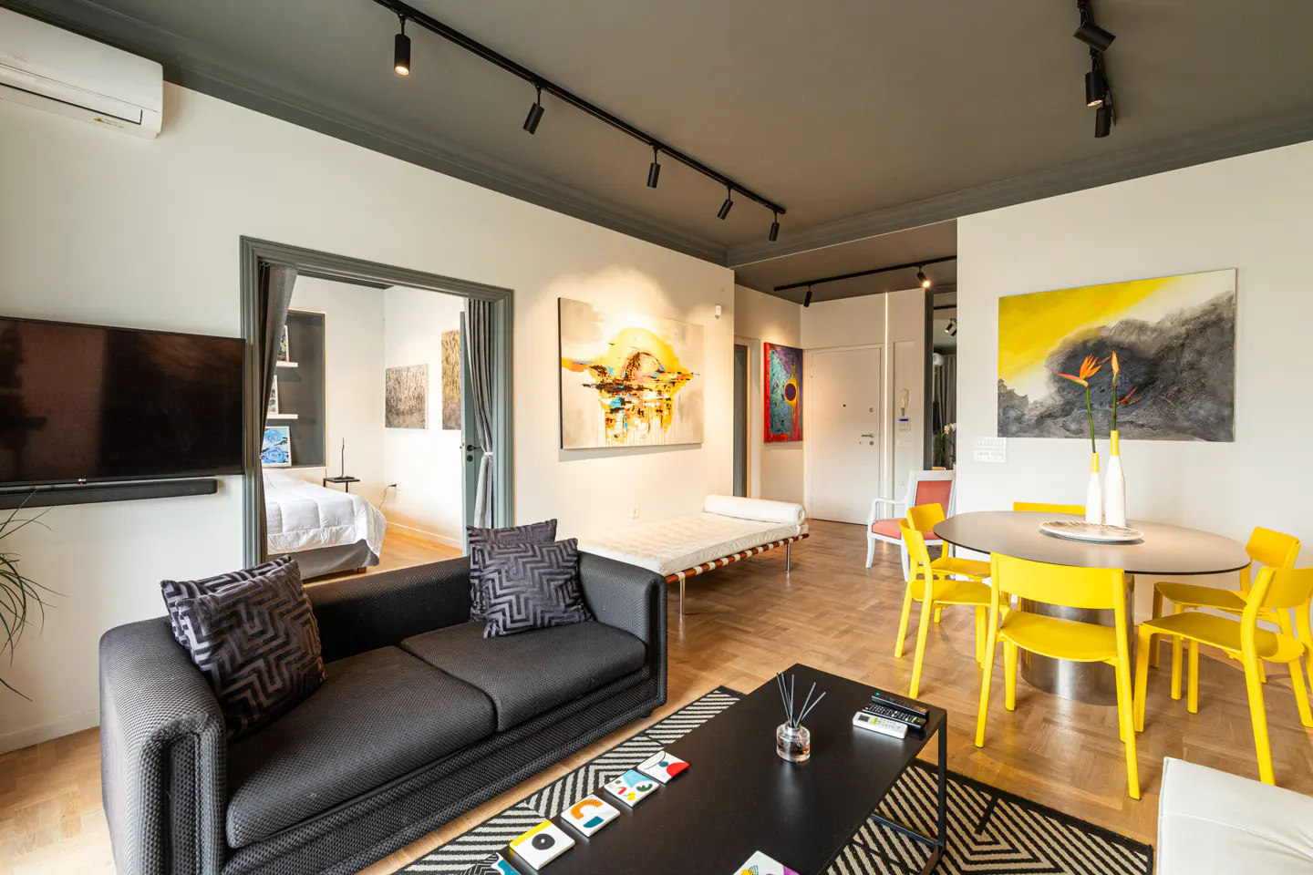 A modern living room with a black sofa, yellow chairs around a table, and abstract art on the walls. A bedroom is visible through an open doorway.