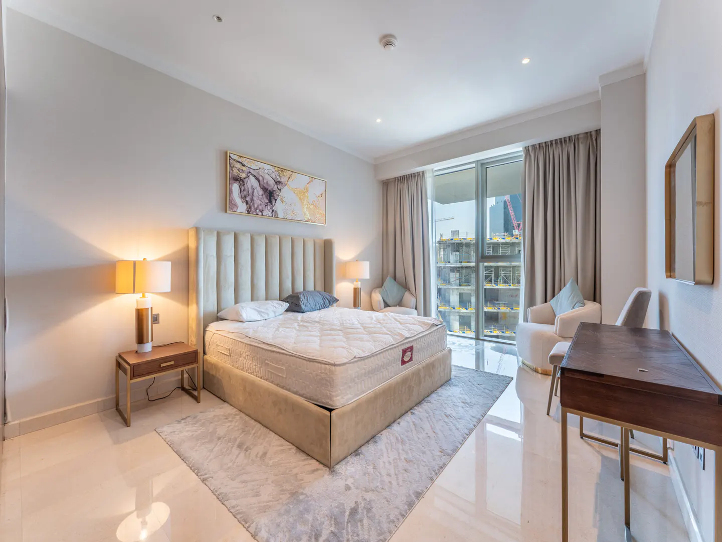 A modern bedroom with a beige bed, a tall headboard, and a desk. A large window shows a city view.