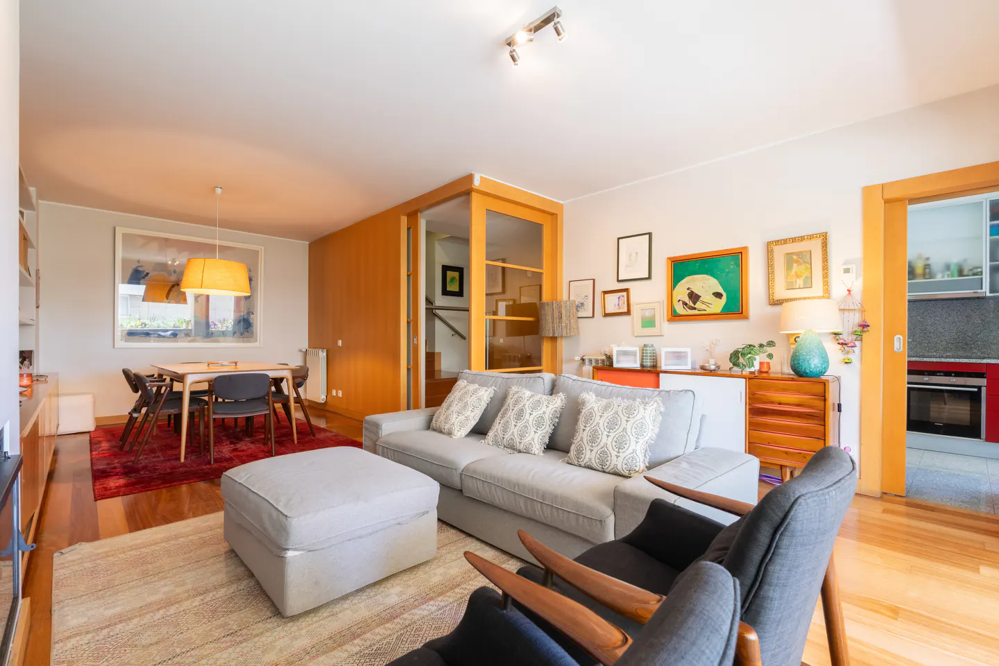 Living room with gray sofa, ottoman, and chairs. Dining table with chairs, wood floors, and art on the walls.