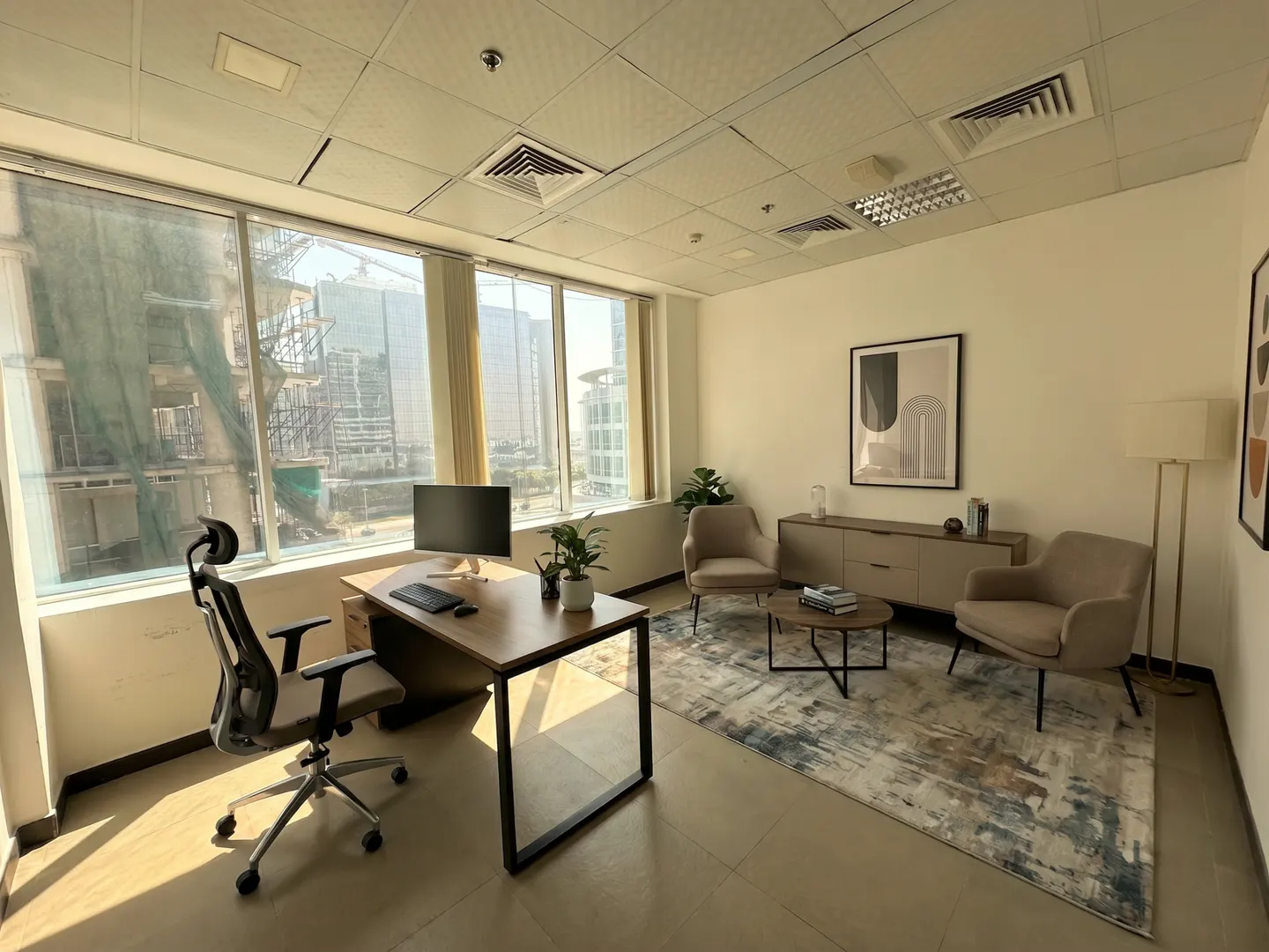 Modern office with a wooden desk, ergonomic chair, two beige armchairs, a coffee table, and city views through windows.