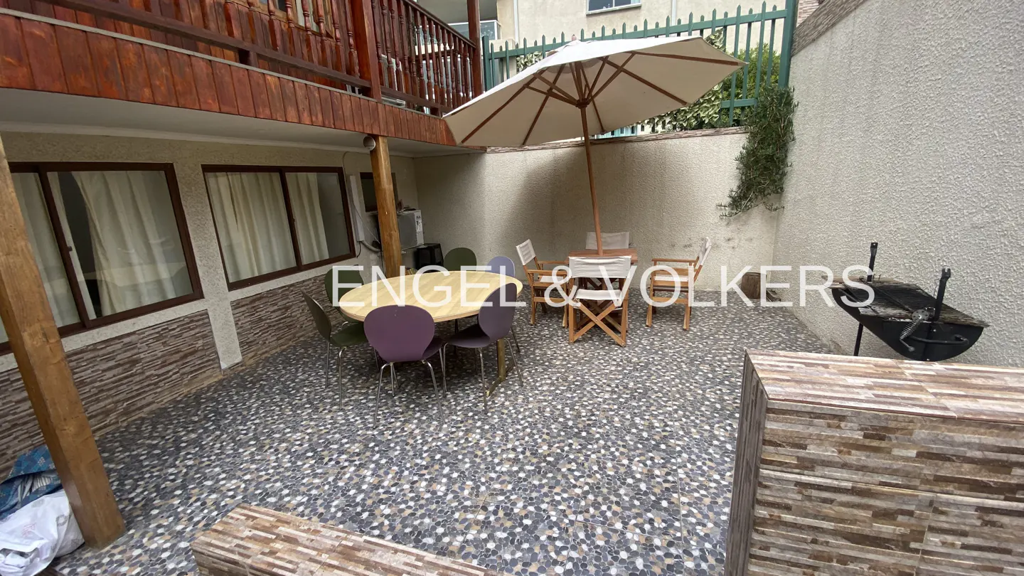 Outdoor patio with stone floor, table, chairs, umbrella, and grill. Balcony above. Engel & Volkers logo.