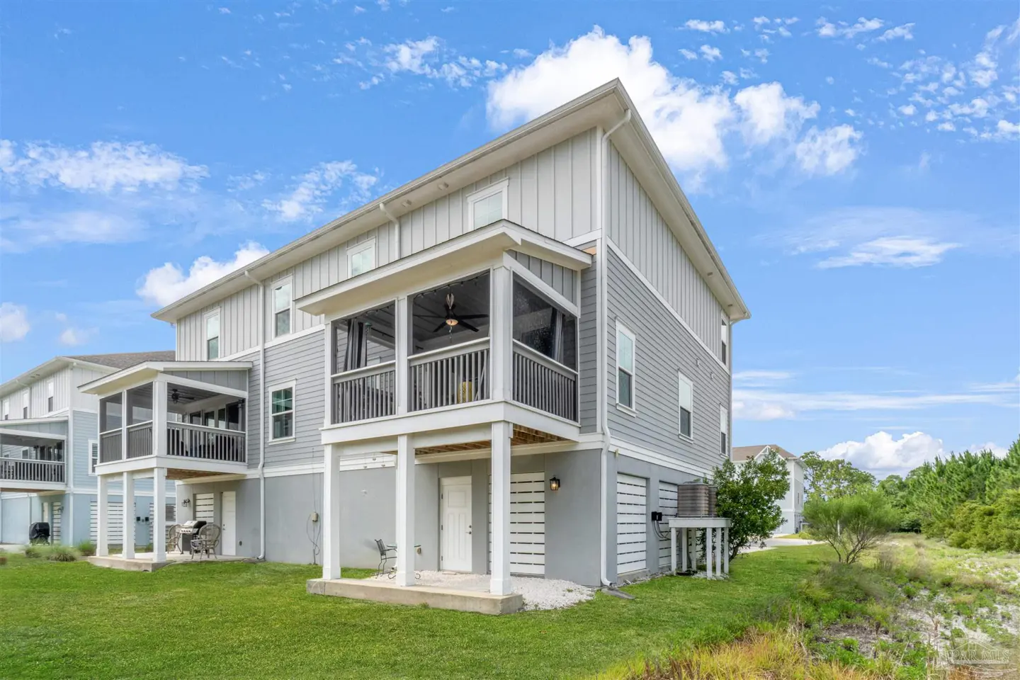 Rear view of a two-story gray house with screened porches, white trim, and green lawn under a blue sky.