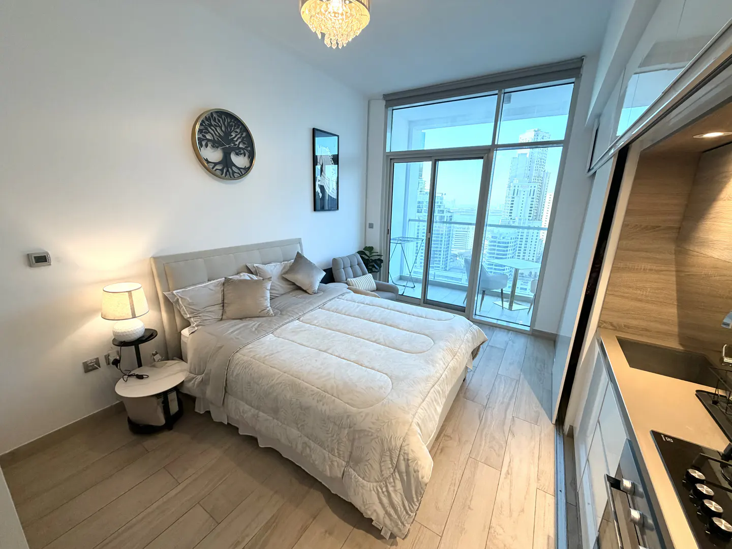 Modern studio apartment with a queen bed, bedside lamp, tree wall art, and floor-to-ceiling windows overlooking a city view.