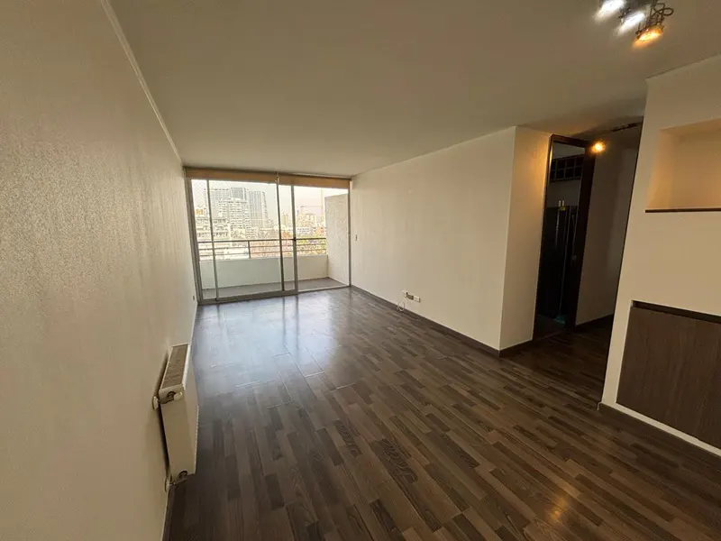 Bright, empty living room with dark wood floors, white walls, and sliding glass doors to a balcony with a city view.