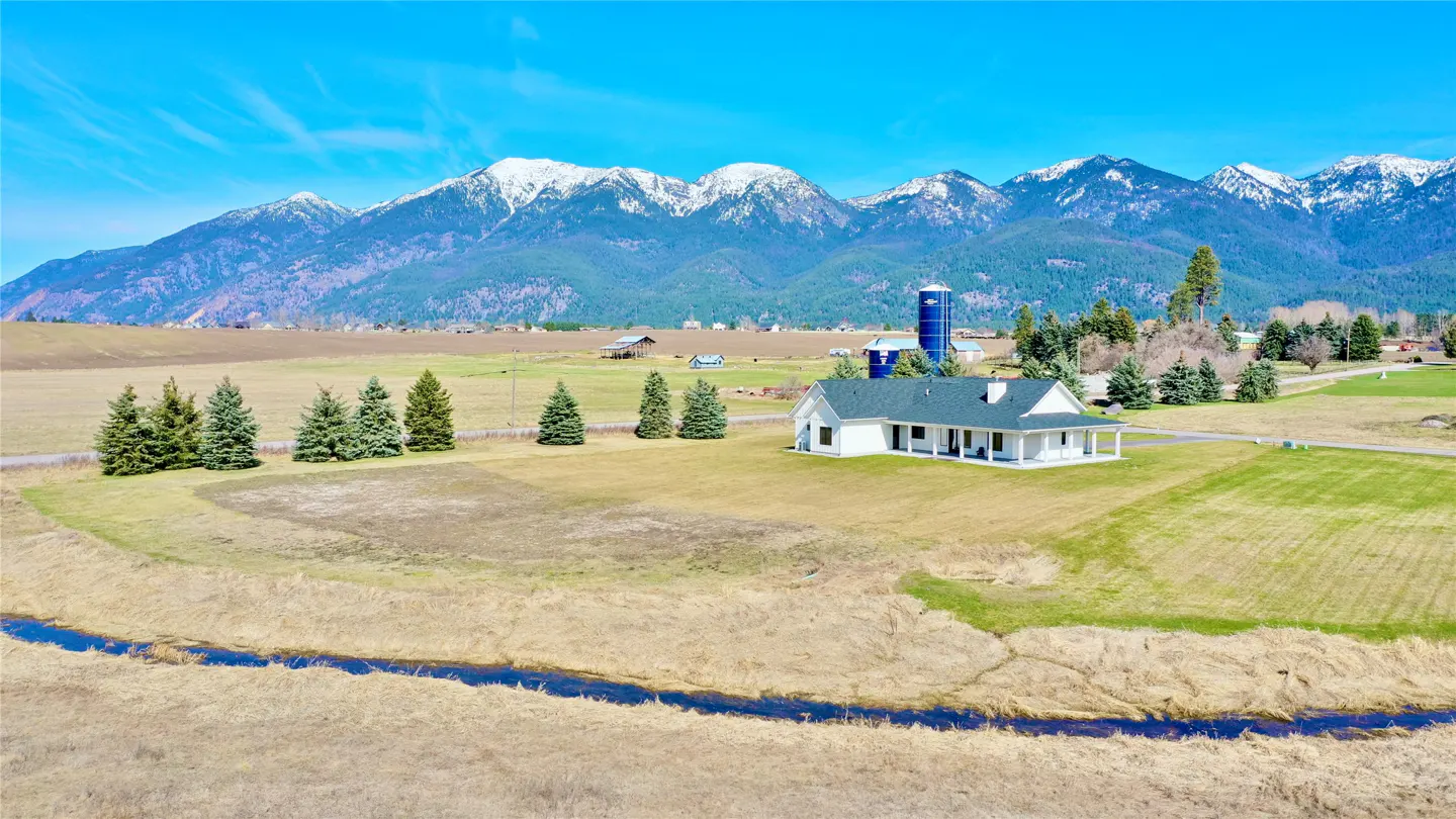 A white farmhouse with a green roof sits on a large lawn with snow-capped mountains in the background. A blue silo is visible.