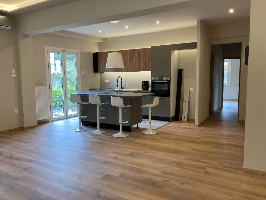 Open-concept kitchen with wood floors, gray island with white stools, and stainless steel appliances. A doorway leads to another room.